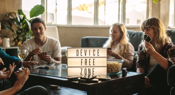 A group of friends sitting together playing cards, with a sign on the table reading 'Device Free Zone' and smartphones placed aside.