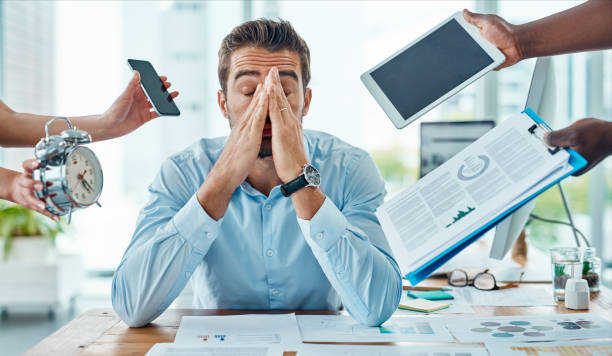 Overwhelmed businessman with hands on face, surrounded by digital devices, paperwork, and a clock, showing the effect of stress on focus and performance.