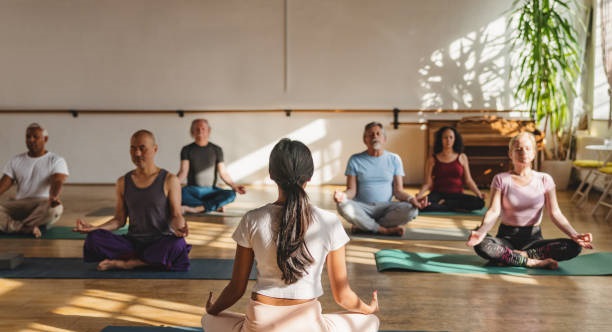 A diverse group of people practicing meditation in a peaceful yoga studio, guided by an instructor. They sit cross-legged, focusing on mindfulness and breathwork.