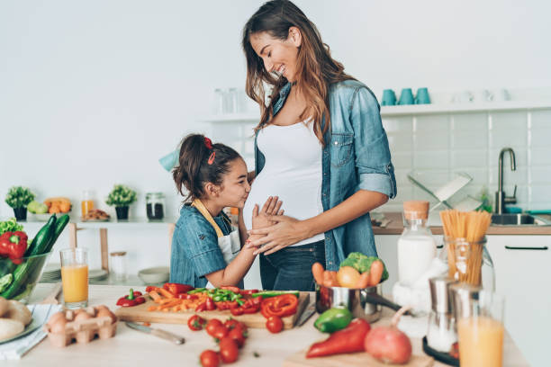 A pregnant mother smiling as her young daughter touches her belly in a kitchen filled with fresh vegetables, symbolizing healthy pregnancy nutrition.