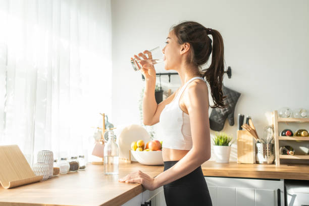 A woman drinking a glass of water in a bright kitchen, emphasizing the importance of hydration for overall health and well-being.