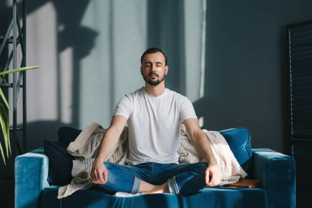 A man sitting cross-legged on a blue couch with his eyes closed, meditating in a peaceful indoor setting. Sunlight streams in through a nearby window, casting soft shadows on the wall. This image represents how to meditate in a comfortable and relaxing home environment.