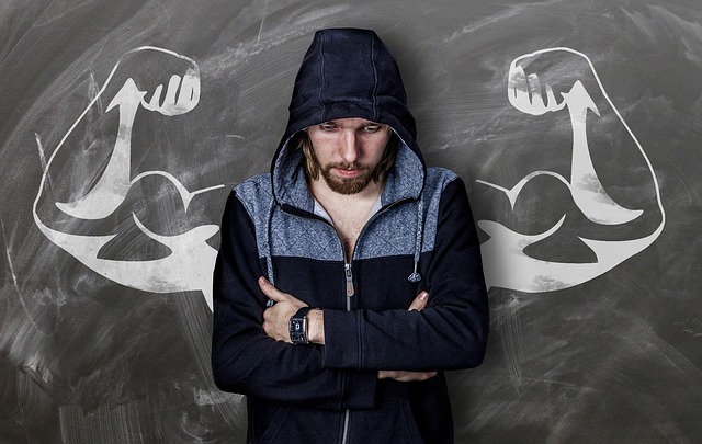 A skinny man in a hoodie stands with crossed arms, looking down, with muscular arms drawn behind him on a chalkboard, symbolizing strength and confidence.