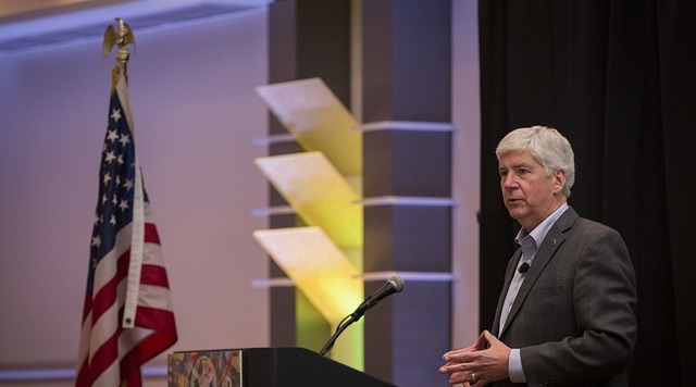 A professional speaker delivering a speech at a conference with a microphone and an American flag in the background.