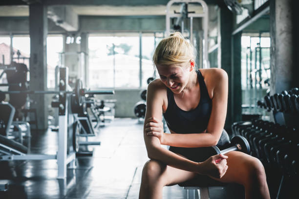 A woman sitting on a gym bench holding her arm in discomfort, highlighting the importance of learning how to prevent injuries while exercising.