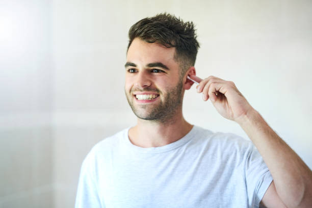 A smiling man using a cotton swab to clean his ear, practicing ear hygiene in a bright bathroom setting.