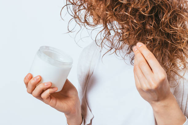 A woman holding a jar of hair conditioner while applying it to her curly hair, following the best way to condition your hair for hydration and smoothness.