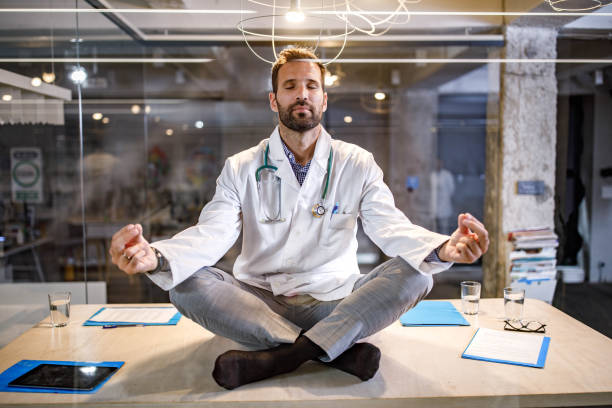 A doctor in a white coat sitting cross-legged on a desk, meditating with a calm expression, representing the benefits of Yoga Poses for Pain Relief.