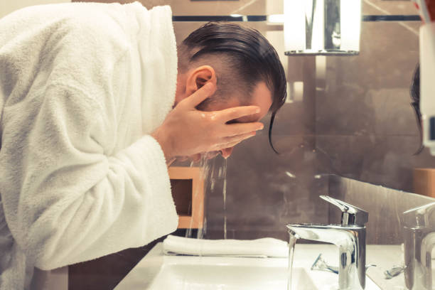 Man in a white robe washing his face at a bathroom sink.