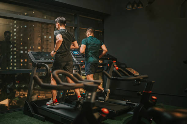 Two men engaged in a night workout, running on treadmills inside a dimly lit gym. The large glass windows reveal a cityscape with glowing lights, adding to the modern and focused workout atmosphere.