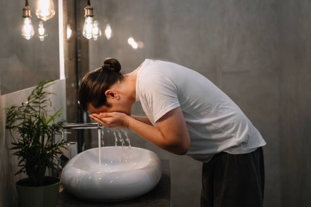 A man with long hair tied back washing his face at a modern sink, emphasizing the importance of washing your face before bed for clear and healthy skin.