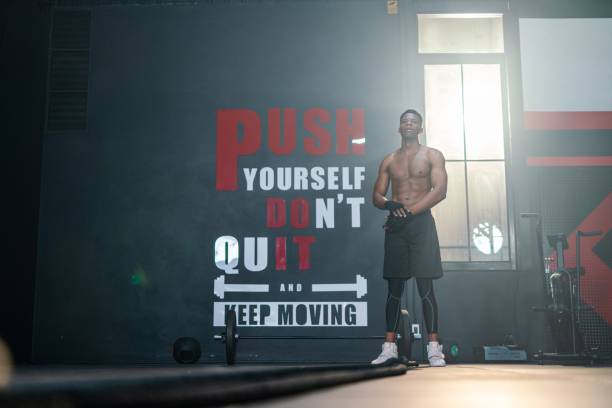 A fit, muscular man in a gym standing confidently in front of an inspirational workout quote on the wall, promoting motivation and fitness consistency.