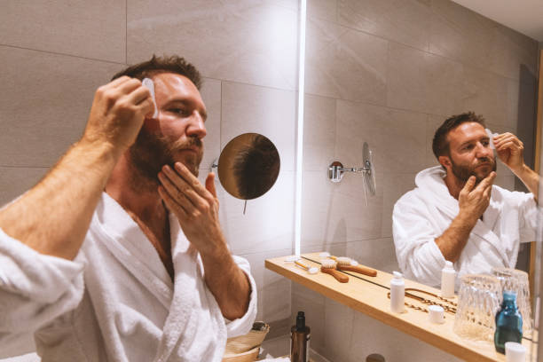 Man in a bathrobe applying a nighttime skincare routine for skin repair in front of a bathroom mirror, using skincare products for hydration and rejuvenation.