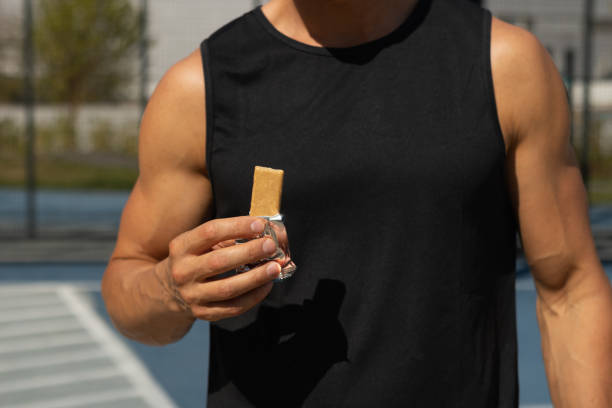 A muscular athlete in a black tank top holding a high-protein snack bar outdoors, highlighting a nutritious post-workout energy boost.