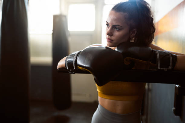 A focused female boxer in gloves resting on the ropes, symbolizing the dedication needed to improve stamina and endurance through consistent training.