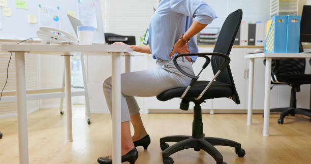 A woman sitting at her office desk holding her lower back in pain due to poor posture, highlighting the effects of improper sitting habits.