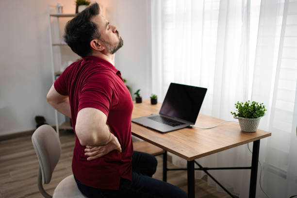 A man sitting at a desk with his hands on his lower back, stretching uncomfortably, showing the effects of poor posture and how to Alleviate Pain from prolonged sitting.