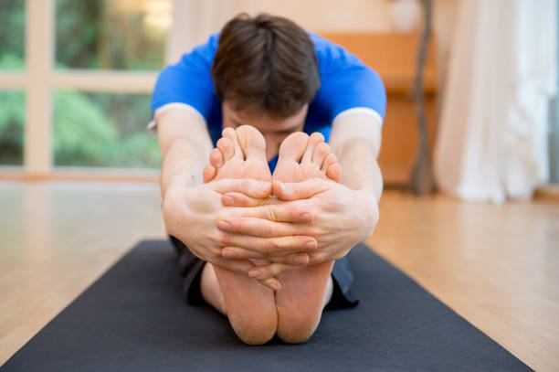 A man practicing Seated Forward Bend yoga pose, bending forward over extended legs while holding his feet, helping with hamstring flexibility and lower back pain relief.